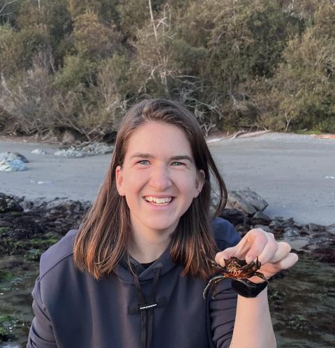 Molly Urtz, smiling, holds up a crab to show the camera, with rocks, then sand, and trees in the background
